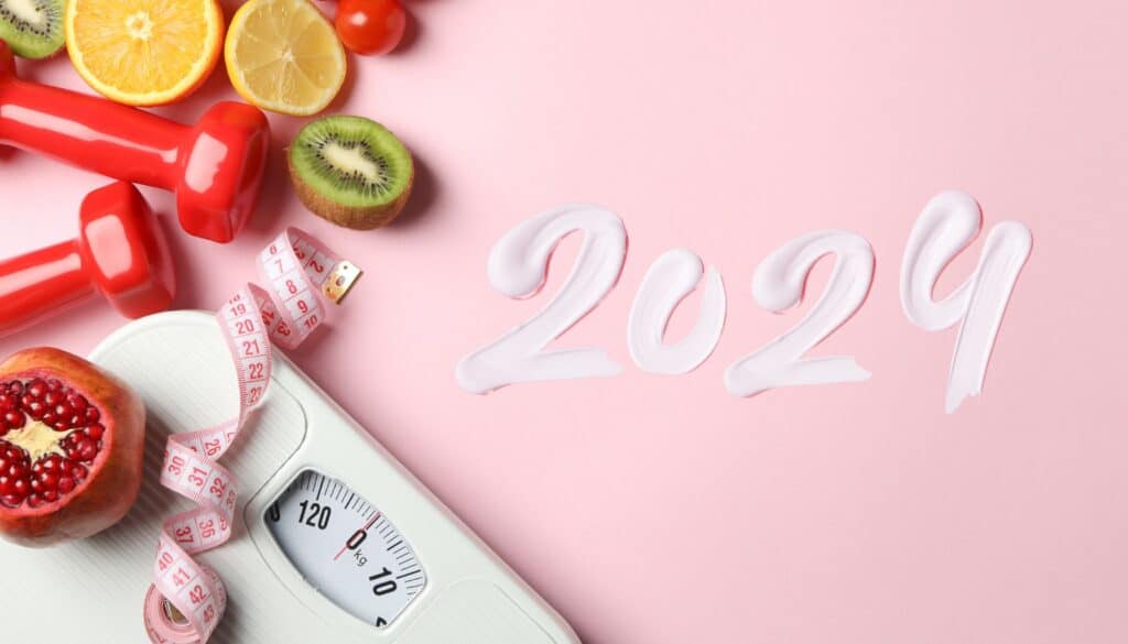 Image de préparation pour 2024 avec des haltères rouges, fruits frais, ruban de mesure et balance, illustrant l'article de Catlion sur la planification d'objectifs de santé et de fitness pour une année réussie.